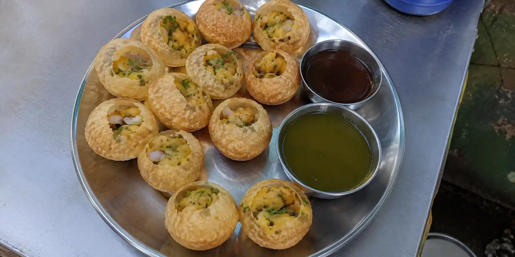 A large brass plate with crispy golgappa puris filled with ragda and potato, bowls of green spicy mint water and brown imli chutney, with sev and coriander scattered on top on a wooden table.