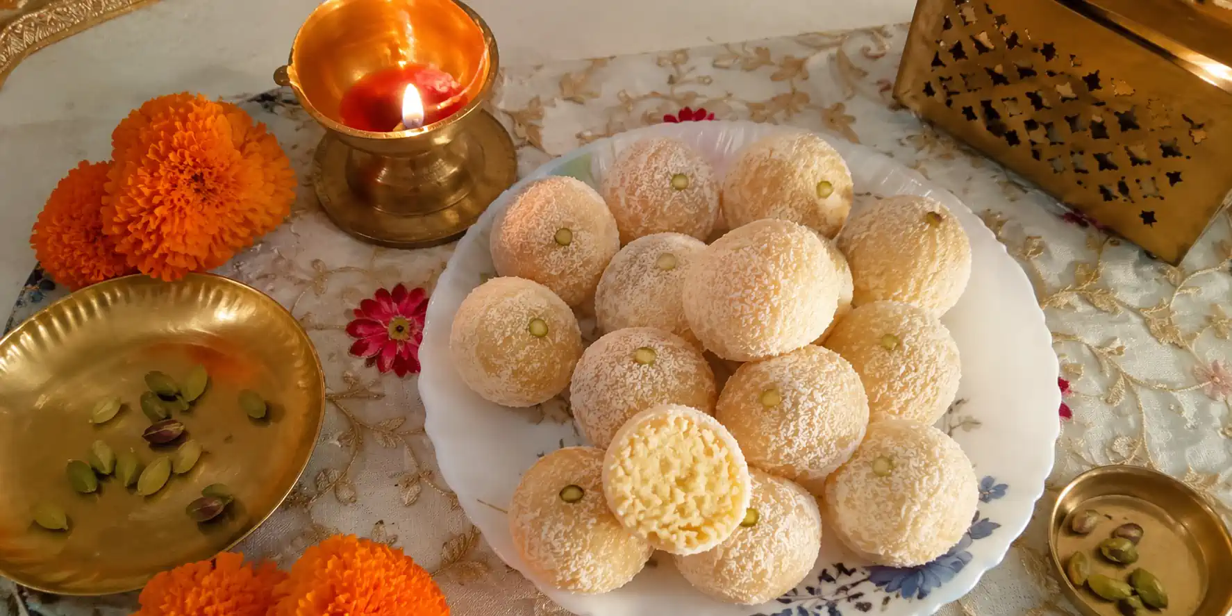 A plate of golden-white milkmaid ladoo, coated in desiccated coconut, arranged in a festive setting with a diya and marigold flowers.