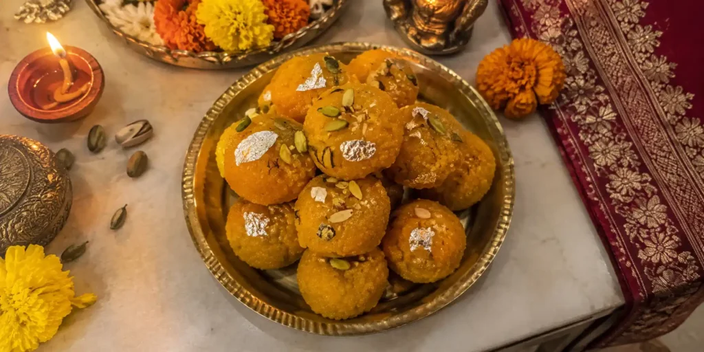 A brass plate with golden-orange motichoor ladoos garnished with pistachio slivers and silver foil, with a diya and marigold flowers on a festive background.